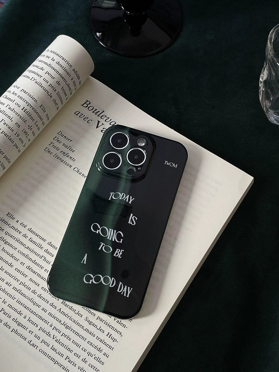 A smartphone encased in a "Good Day Quote iPhone 16 Pro Max Case" with a sleek dark green design and the motivational phrase "Today is going to be a good day" rests on an open book. In the background, you can partially see a glass and a black object.