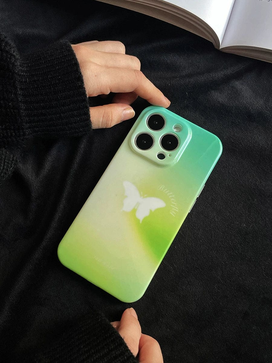 Close-up of a person holding the Butterfly Bliss Gradient iPhone 16 Pro Max Case with its soothing green and butterfly design, placed on a dark fabric surface with an open book in the background.