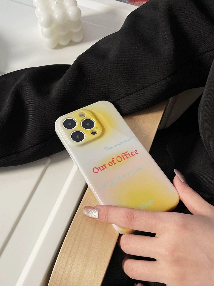 A person is holding the Out of Office iPhone 16 Pro Max Case with a stylish gradient yellow design. The case, featuring text that reads "Out of Office" over black fabric, has a bubble-like decorative item visible in the background.