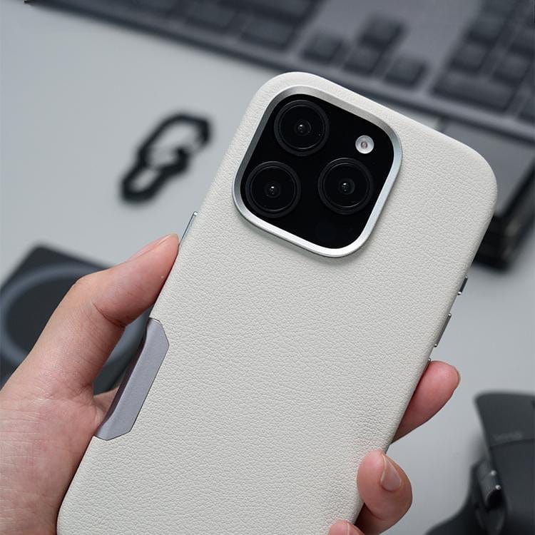 The smartphone in the hand is fitted with the iPhone 16 Pro Max Leather Case, featuring a white textured design and showcasing a triple camera setup. This case is crafted from premium leather for a luxurious feel and boasts MagSafe compatibility, allowing for seamless charging and accessory integration.