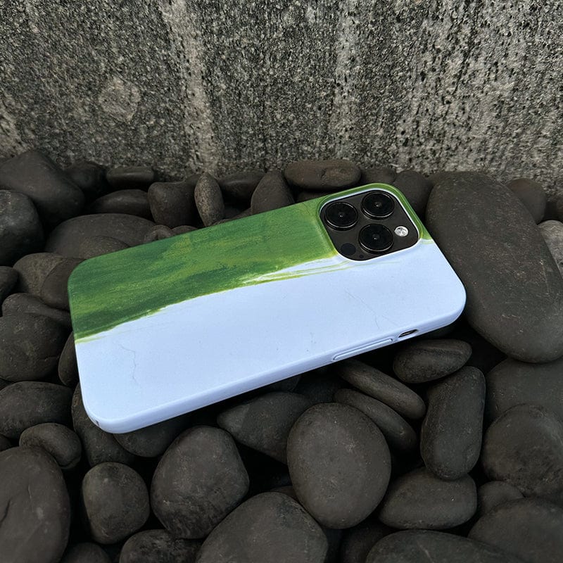 A smartphone with a Minimalist Green Brushstroke iPhone 16 Pro Max Case, featuring an artistic abstract design in a green and light blue gradient, lies face down on a bed of smooth, dark gray stones, with a textured, weathered concrete wall in the background. The phone's camera lenses are visible in the upper corner.