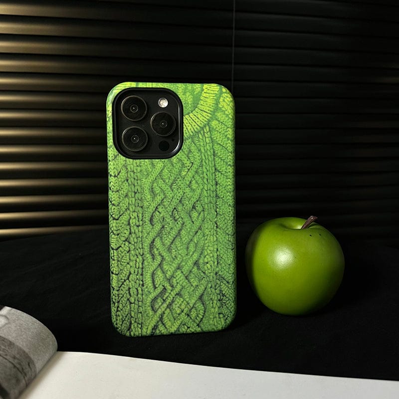 A smartphone with a Cozy Knitted Sweater iPhone 16 Pro Max case featuring a textured green design rests on a black surface next to a green apple. The background displays horizontal dark-striped lines, and part of a white document or piece of paper protrudes from the left corner.