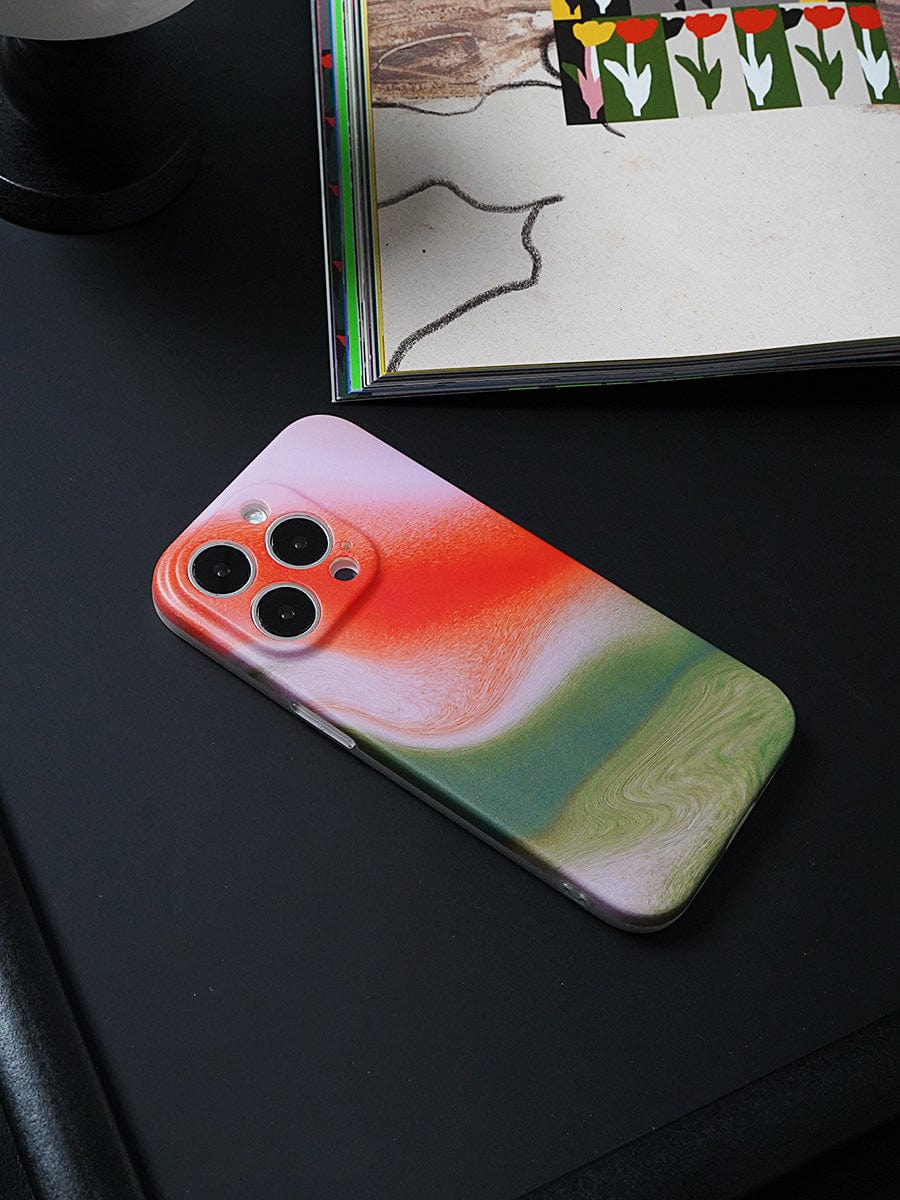 A smartphone with a Sunset Swirl iPhone 16 Pro Max case, featuring a stylish abstract gradient design in red, white, and green hues, lies on a dark surface next to an open book with tulip illustrations.