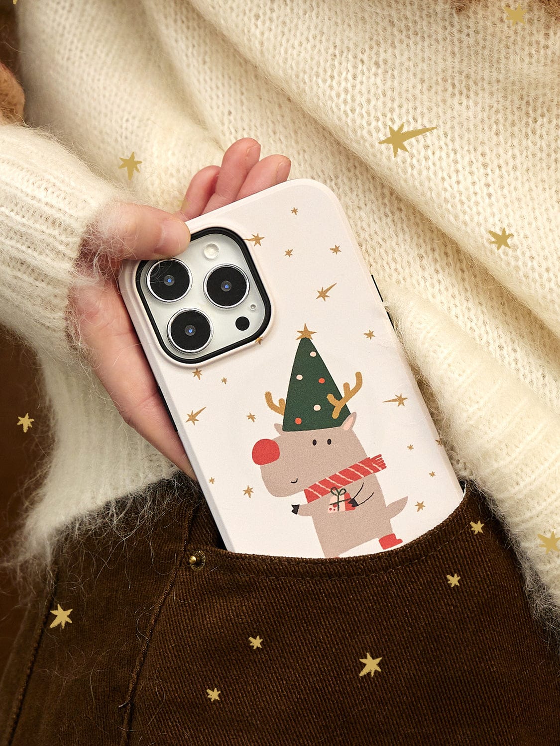 A hand in a sweater and brown pants holds the Rudolph Joy MagSafe iPhone 16 Pro Max Case, featuring a festive reindeer design decorated with stars.