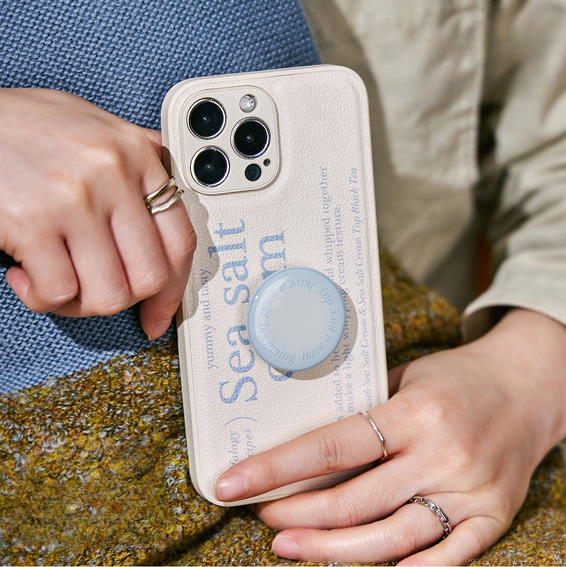 Person holding an iPhone with a sleek Sea Salt Cream MagSafe iPhone 16 Pro Max Case, featuring a minimalist textured design, text, a blue PopSocket, and a triple-camera setup.