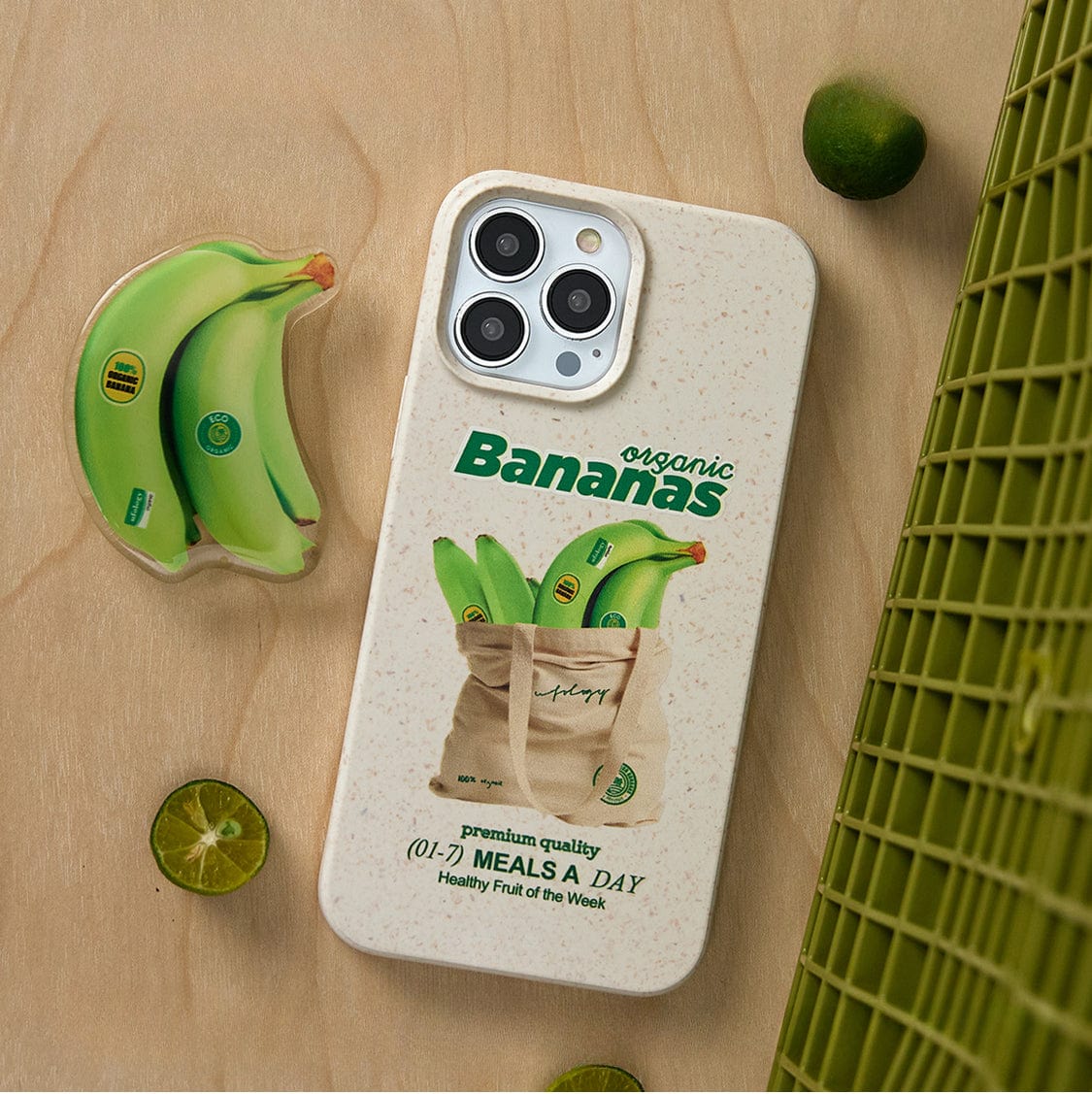 A smartphone encased in an Organic Bananas MagSafe iPhone 16 Pro Max case, featuring a quirky banana graphic and green "organic Bananas" text, rests on a wooden surface near a tote bag. Alongside it are a banana-shaped sticker, green grid material, and sliced lime.