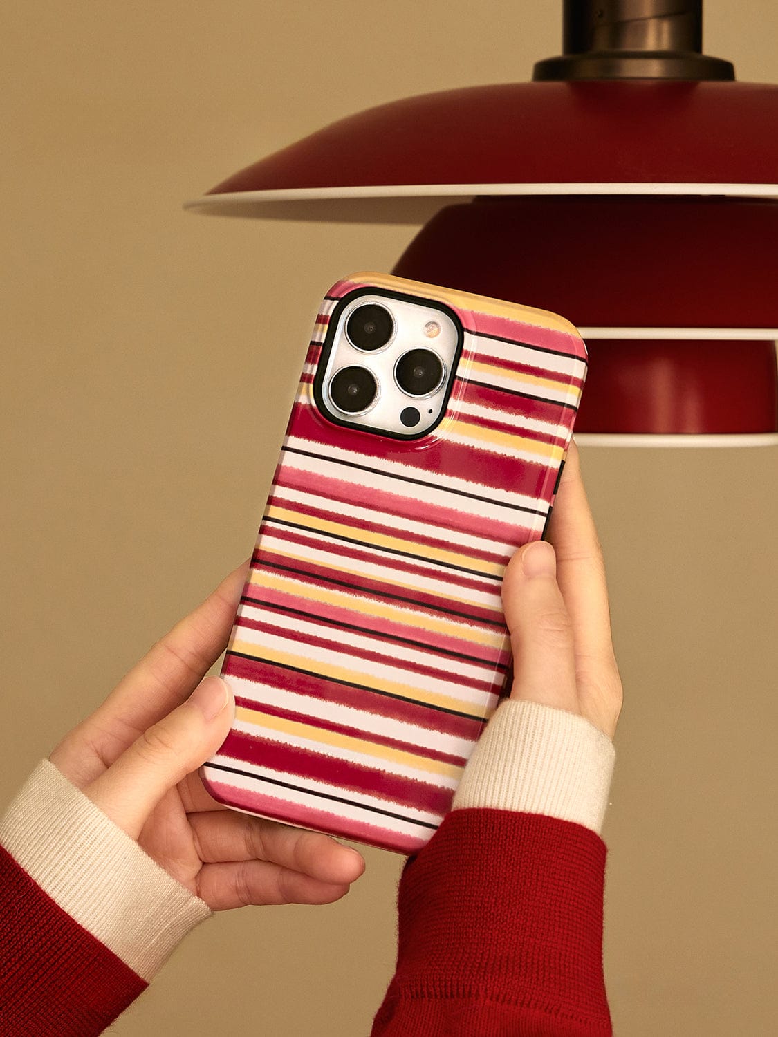 A person wearing a red and white sweater holds a smartphone with an Autumn Stripes MagSafe iPhone 16 Pro Max Case, featuring a cozy mug design with a stand. The phone is positioned against an orange lamp shade in the background.