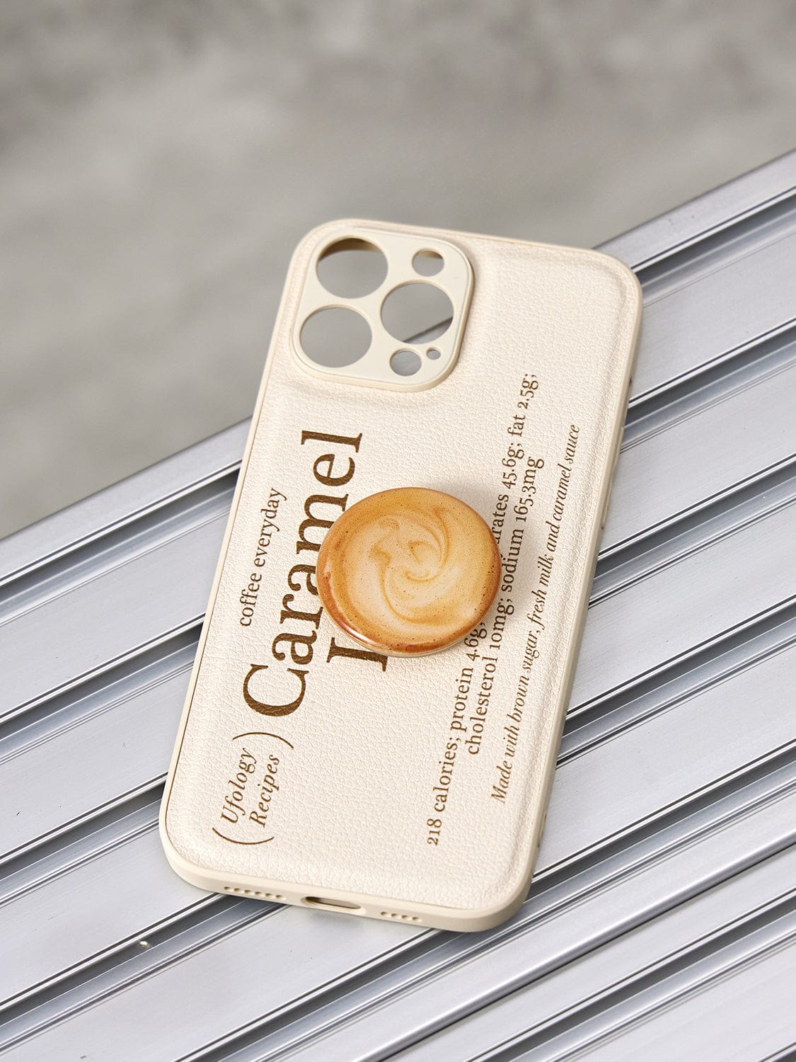 A Caramel Latte MagSafe iPhone 16 Pro Max case, featuring a coffee-inspired design and an integrated cup stand, lies on a metallic surface. The light-colored protective cover showcases text about caramel and includes a circular PopSocket adorned with a caramel swirl pattern for easy grip.