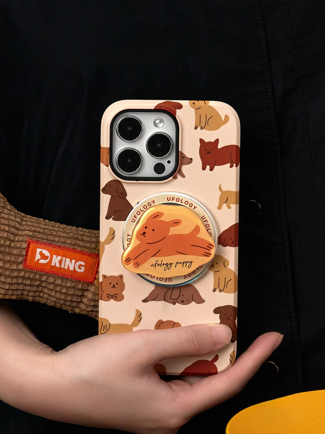 A hand holds a Puppy Playtime MagSafe iPhone 16 Pro Max Case, featuring a variety of cartoon dogs in beige and an integrated pop socket with another cartoon dog. The case is complemented by a "D King" label on a brown strap, providing both style and all-inclusive protection.