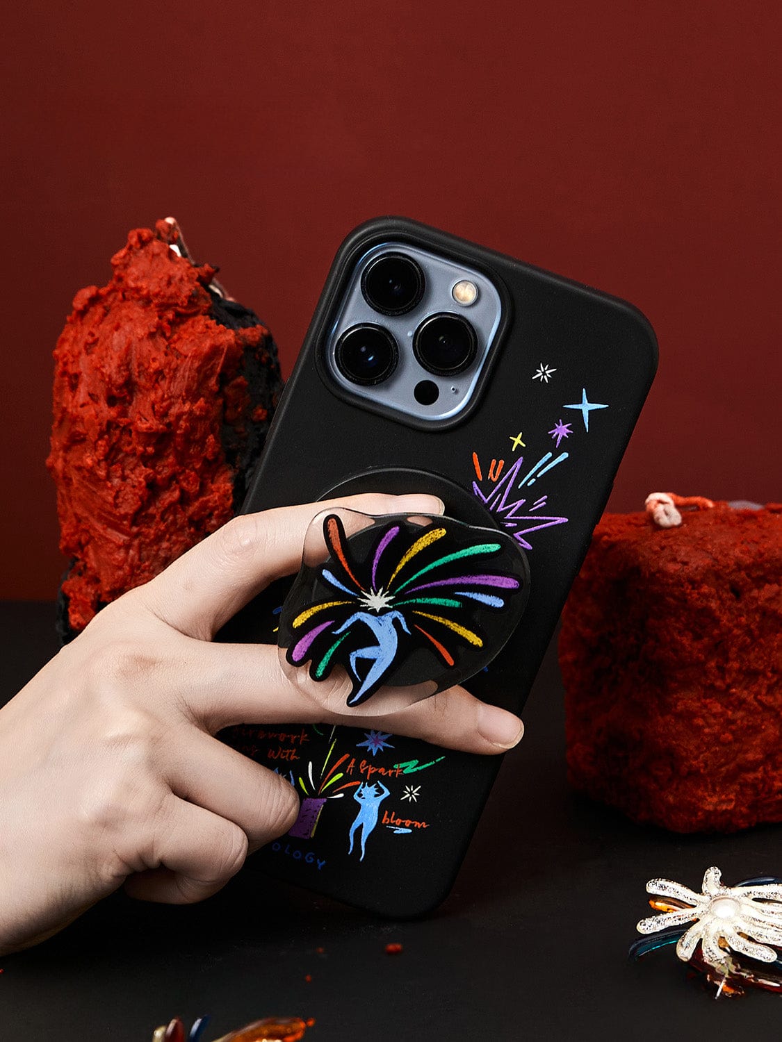 A hand holding an iPhone 16 Pro Max encased in a Firework Fantasy MagSafe Case, showcasing its vibrant celebration design. The phone features a stand adorned with a multi-colored spiral pattern on the back. Red textured objects are visible in the background against a dark surface.