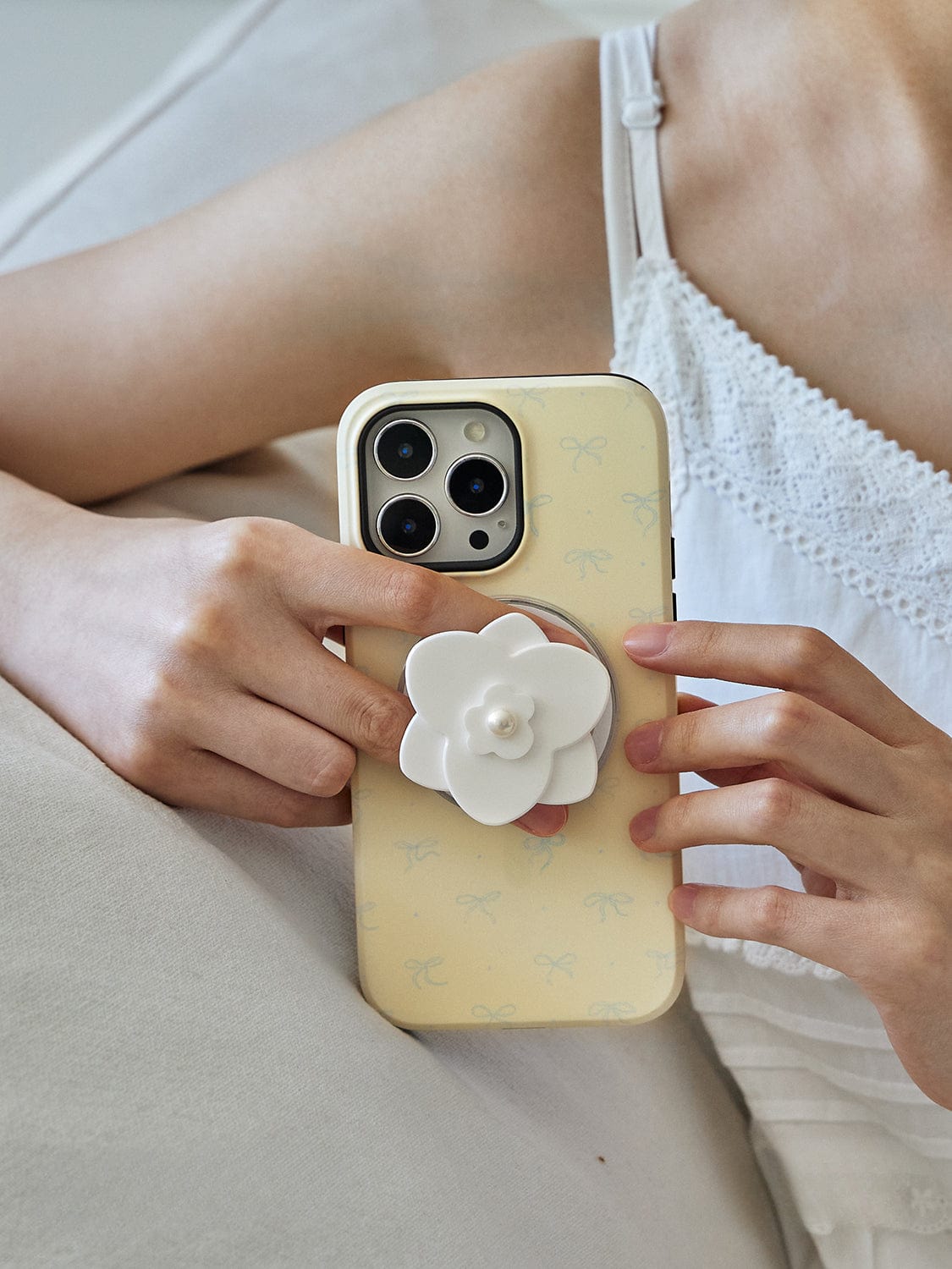 A person in a white lace top holds an iPhone 16 Pro Max with an Elegant Blossom MagSafe Case that features a chic floral design and is topped with a pearl flower stand.