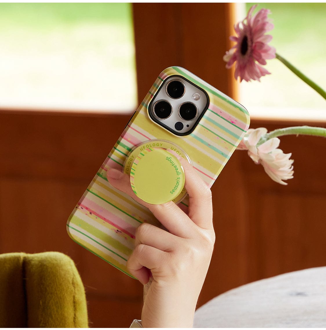 A hand holds the Festive Stripes MagSafe iPhone 16 Pro Max case, which features colorful stripes in pastel green, yellow, and pink, along with a lime green PopSocket. The scene is set against a backdrop of wooden furniture and a pink daisy in a vase.