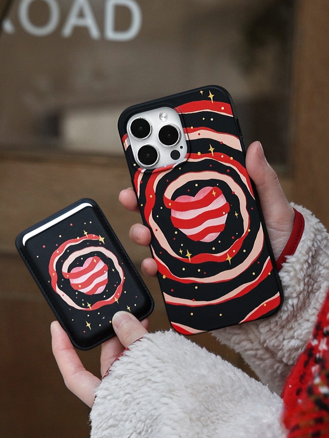 A person holding the Galaxy Heart MagSafe iPhone 16 Pro Max case and a matching cardholder. Both items feature an artistic design with swirling red, black, and white patterns, depicting a planet and stars in a space-themed motif. The person wears a fluffy sweater, with a blurred background visible behind them.