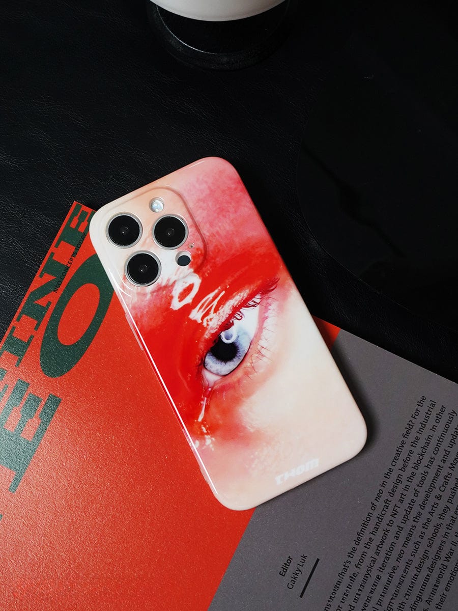 The Red Eye Art iPhone 16 Pro Max Case, designed for bold artistic expression, features a close-up image of an eye with dramatic red makeup. It is placed on top of a red and black book or magazine, partially obscuring the text on the cover.