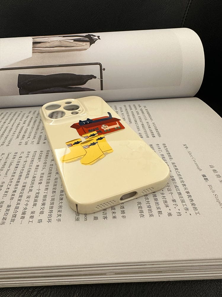 A smartphone with a Cute Cat in Rain Boots iPhone 16 Pro Max case featuring a playful cartoon design lies on an open Chinese book. The cartoon depicts a red sofa, a small table, a tea set, and two yellow socks. The background is out of focus, highlighting the phone case.