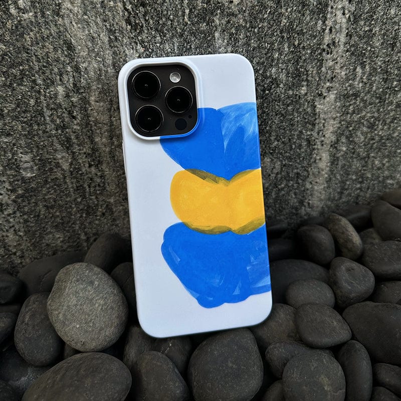 An Abstract Expressionist iPhone 16 Pro Max Case - Bold Blue & Yellow Brushstrokes with a white finish lies on a bed of black pebbles. The case features a hand-painted design of a blue and yellow flower, slightly abstract in style, with large, broad strokes of paint.