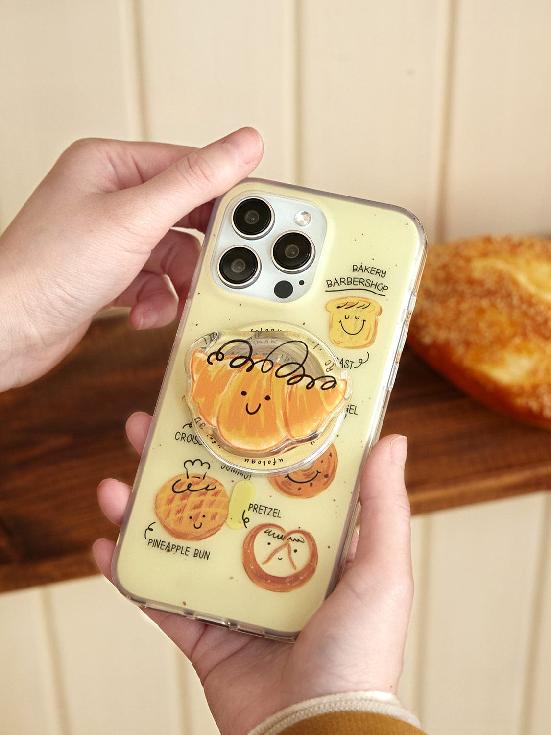 A person holding the Bakery Friends MagSafe iPhone 16 Pro Max Case, which features a clear design with cute cartoon baked goods. The phone case includes a pop socket shaped like a smiling bun. In the background, there is a baked pastry visible on a wooden surface.