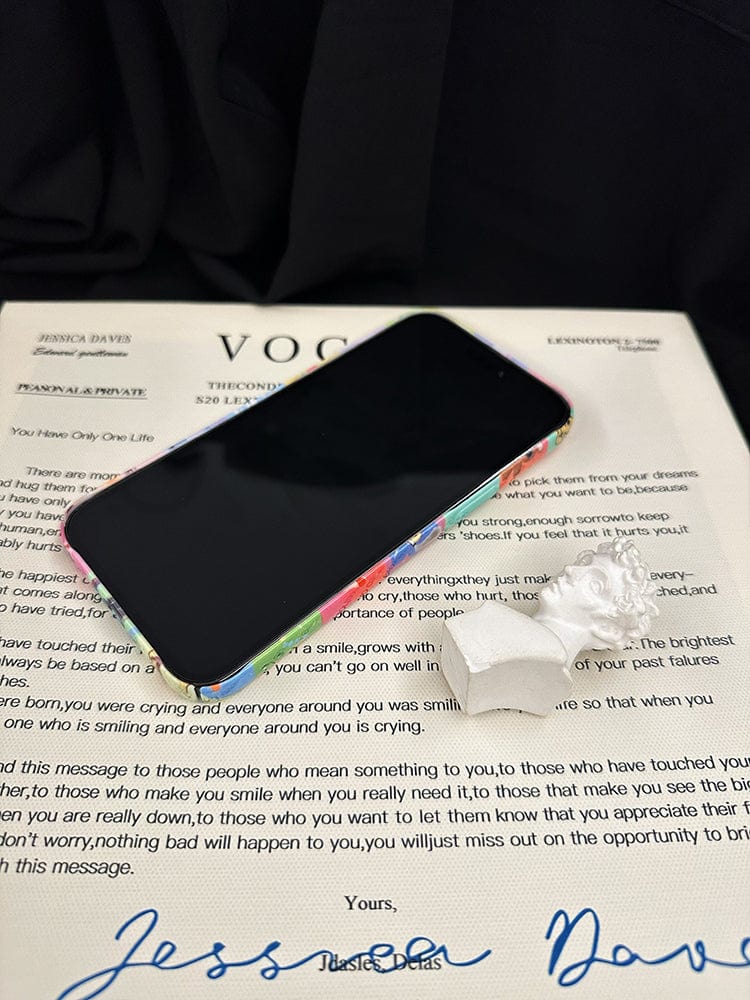 A smartphone encased in a Colorful Kawaii Patchwork iPhone 16 Pro Max Case, a small white ceramic angel, and a paper item are placed on top of a sheet with text and signatures. The background is dark.