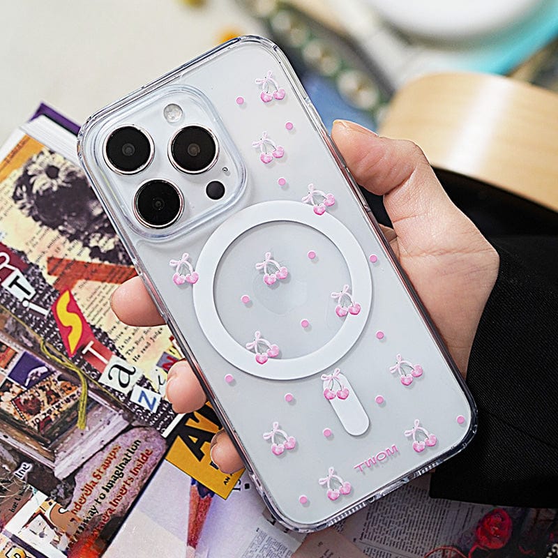 A person holds an iPhone encased in a Cherry Blossom MagSafe iPhone 16 Pro Max Case, adorned with cute 3D cherry illustrations in pink and white. The phone is held over a table with magazines and books in the background.