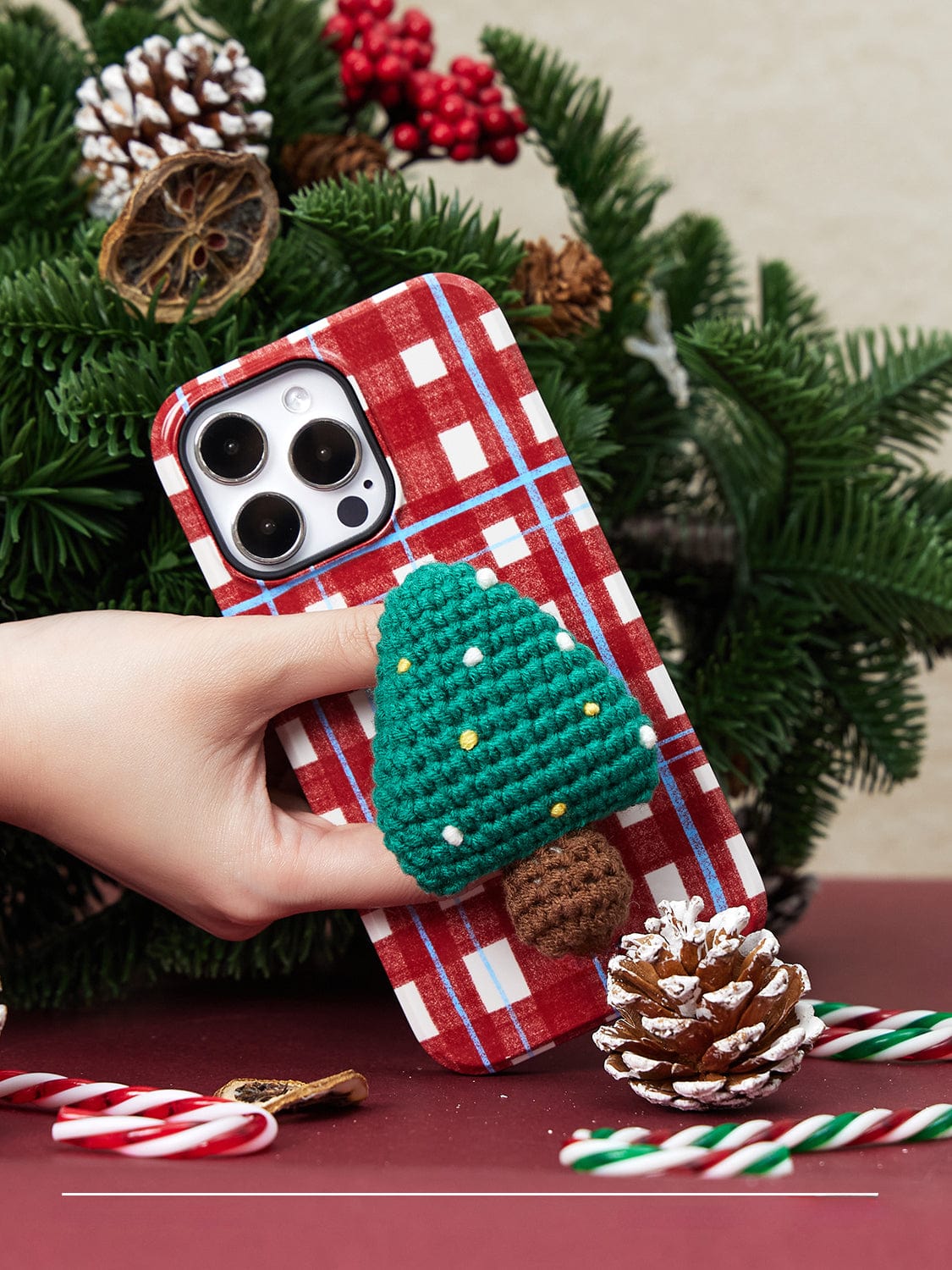 A hand holding the Festive Knitted MagSafe iPhone 16 Pro Max Case with a holiday plaid design and featuring a crocheted green Christmas tree attachment. The background includes festive decorations like pinecones, candy canes, and red berries.