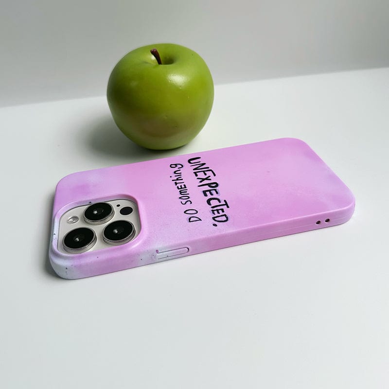 A green apple sits on a white surface next to an Inspirational Pink iPhone 16 Pro Max Case, featuring the 'Do Something Unexpected' quote design. The case is placed face down, showcasing the camera lenses and its ultra-slim, durable protection.