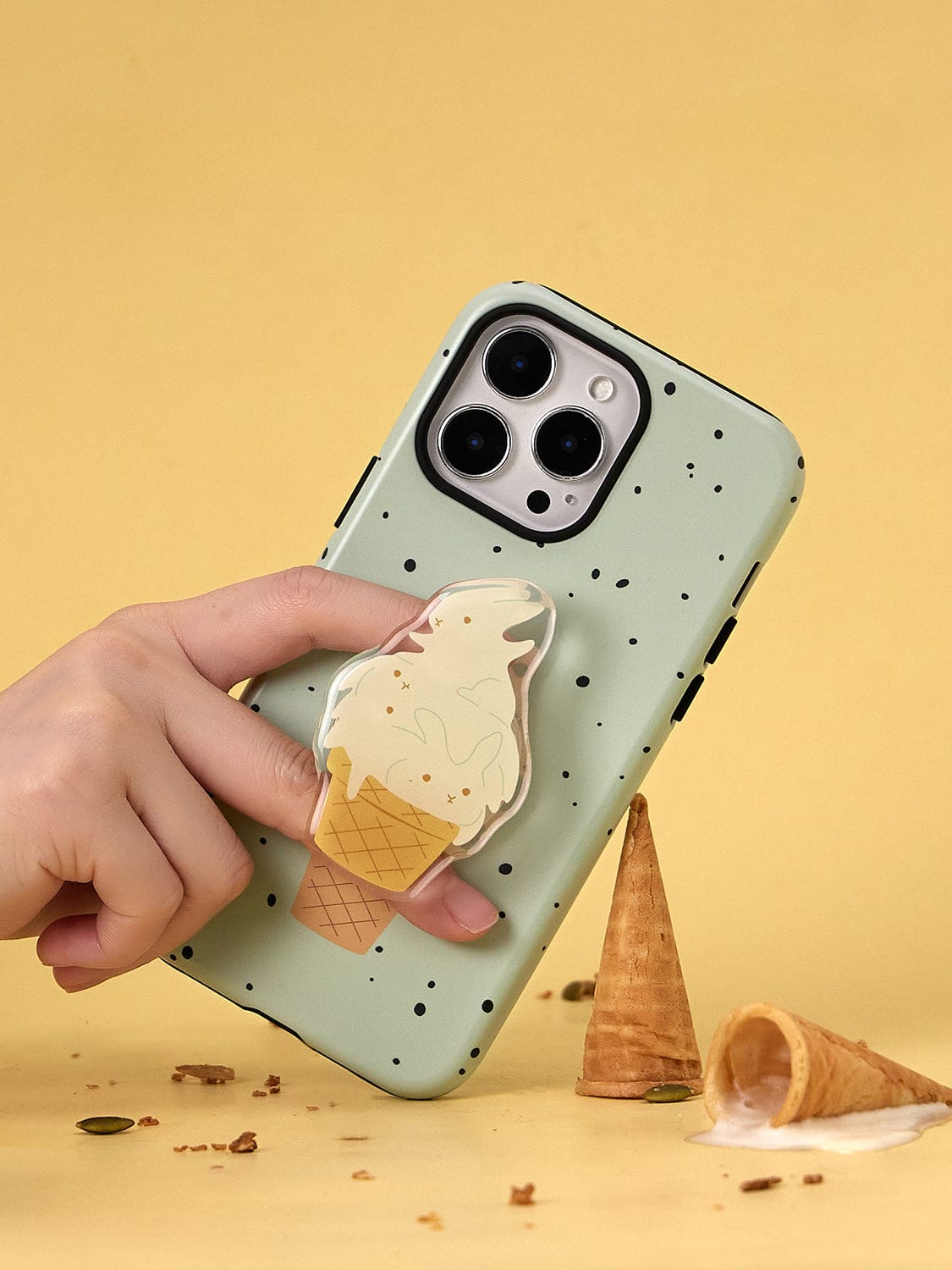 A hand holds the Mint Ice Cream MagSafe iPhone 16 Pro Max Case, showcasing its green color and black-speckled pattern. The case features a grip stand designed like an ice cream cone. The background is yellow with a spilled ice cream cone and small crumbs scattered around, creating the perfect playful setting for this Sweet Treat Design all-inclusive protective cover.