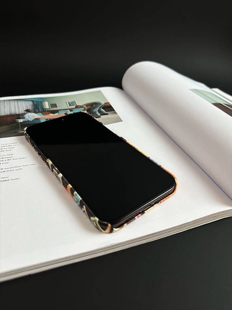 A smartphone adorned with the Vibrant Collage iPhone 16 Pro Max Case, featuring pop art, street style, and retro design elements, is placed on an open book. The pages of the book contain images and text that are not clearly visible. The background is plain and dark, drawing attention to both the phone and the book.