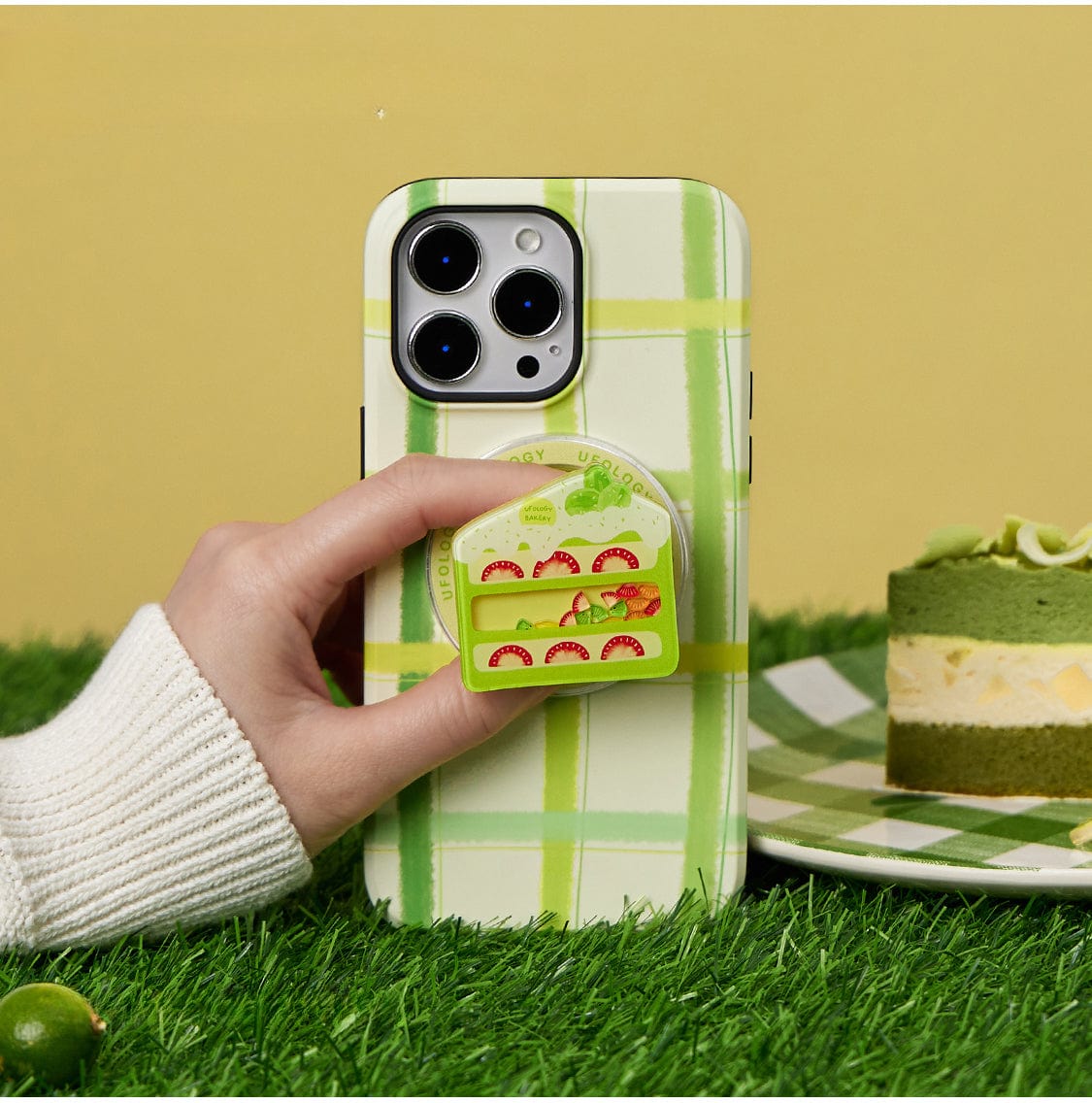 A hand holds a smartphone encased in the Fresh Picnic MagSafe iPhone 16 Pro Max Case, featuring a green plaid design and sandwich stand. A cake-themed pop socket adorns the phone, while a green and yellow layered dessert sits on a matching checkered plate in the background.