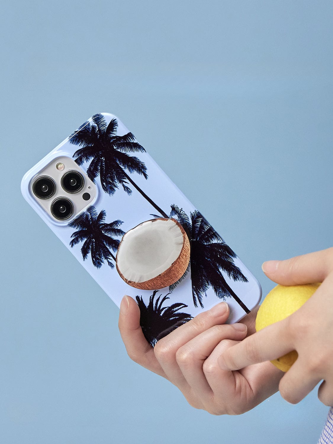 A hand holding the Tropical Escape MagSafe iPhone 16 Pro Max Case, showcasing its palm tree design with a coconut stand on a light blue background. The person's other hand is holding a yellow fruit, possibly a lemon, just below the phone case.