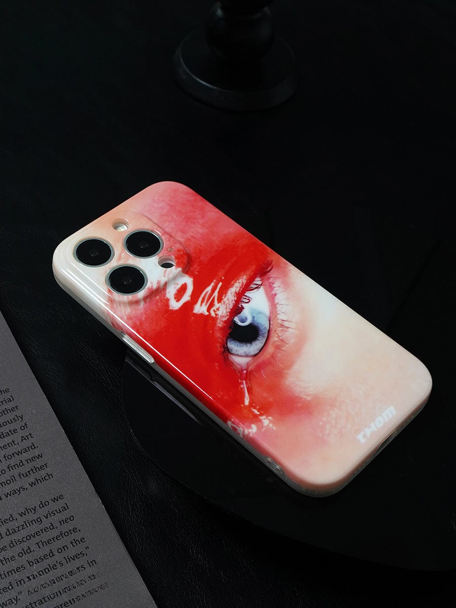 A detailed Red Eye Art iPhone 16 Pro Max Case, showcasing a close-up photograph of a single blue eye surrounded by red makeup, rests on a black surface next to a partially visible open book with text.