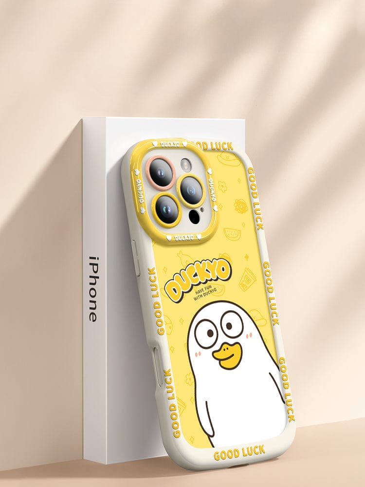 A cute and shockproof iPhone 16 Pro Max Yellow Duck Cartoon Case, labeled "Duckyxo," made of soft liquid silicone, rests on a white iPhone box. Featuring a repeated "Good Luck" message in white around the edges, this slim-fit cover offers anti-yellowing protection and showcases a playful duck design.