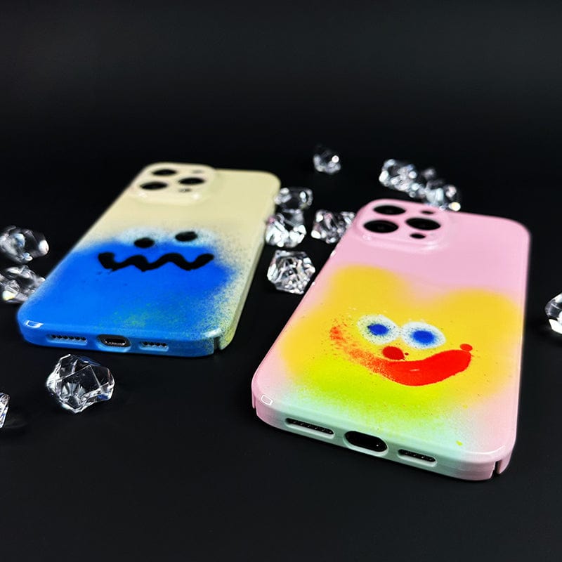 Two cases from the Colorful Spray Art iPhone 16 Pro Max Case Set lie on a black surface, surrounded by scattered clear crystals. The left case features a simple blue face design in blue and white, while the right case showcases a vibrant pink backdrop with a colorful smiling face adorned with a yellow heart and red lips.