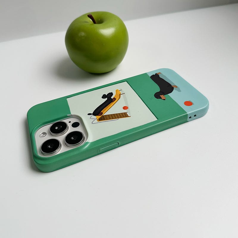 A green iPhone 16 Pro Max with the Playful Dachshund iPhone 16 Pro Max Case - Cute Green & Blue Design is placed on a white surface. The phone case features illustrations of a black dog, a yellow broom, and a small red and white ball. A green apple sits in the background.