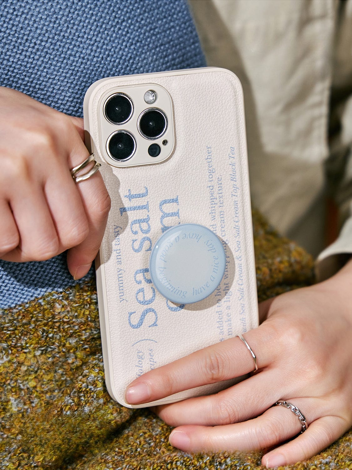 A person holds a Sea Salt Cream MagSafe iPhone 16 Pro Max Case with a minimalist textured design and an inclusive protective cover, featuring text about sea salt and a blue circular phone grip. The person is wearing rings on both hands.