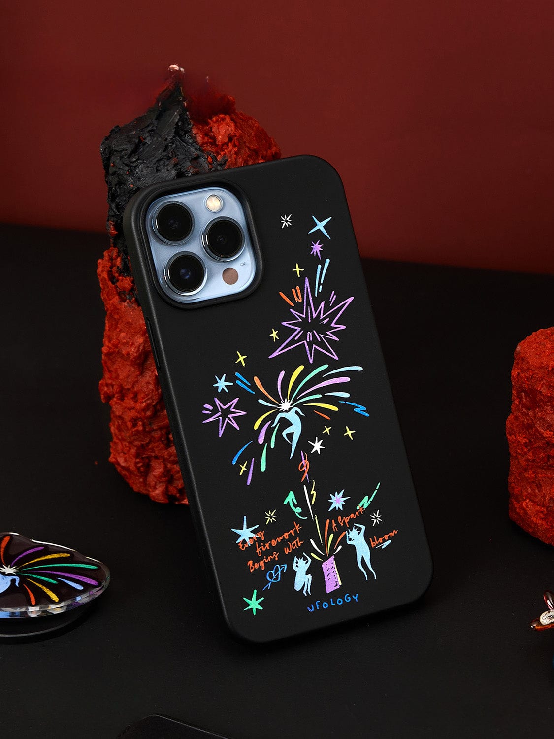 The Firework Fantasy MagSafe iPhone 16 Pro Max Case, featuring vibrant illustrations of aliens, stars, and the phrases "We'll believe it when we see it" and "Get out!", is showcased upright. The background consists of textured red rocks laid upon a dark surface. The brand name "UFOLOGY" is printed at the bottom of the case.