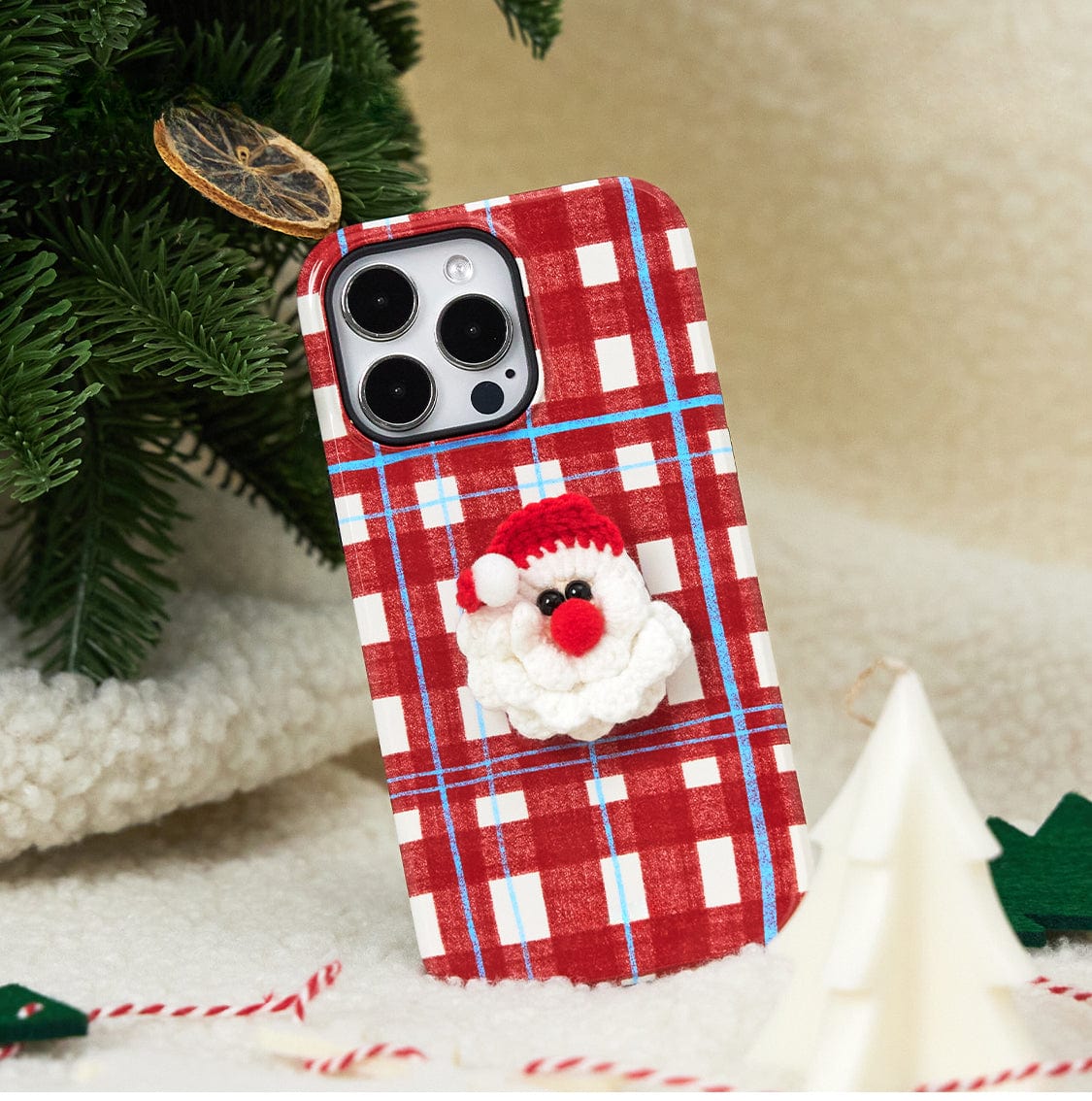 A smartphone with a Festive Knitted MagSafe iPhone 16 Pro Max Case in a holiday plaid design featuring a small, knitted Santa Claus is leaning against a Christmas tree branch. Candy canes and a white tree ornament are situated on a soft, fluffy surface around the phone.