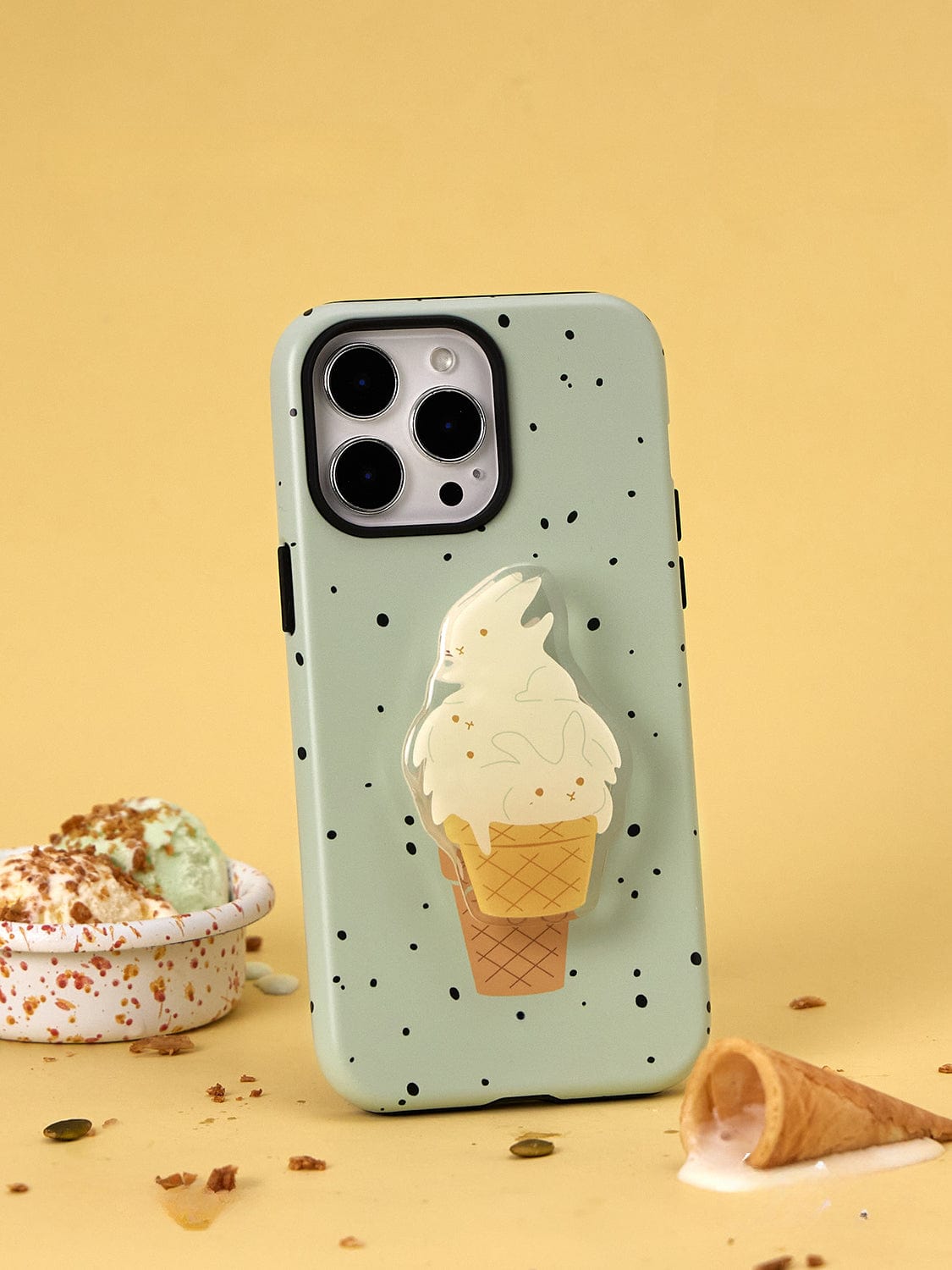 The Mint Ice Cream MagSafe iPhone 16 Pro Max case, featuring a sweet treat design with black speckles and an illustration of a melting ice cream cone, stands upright on a yellow background. Next to it lies a tipped ice cream cone and a bowl of pistachio ice cream with sprinkles.