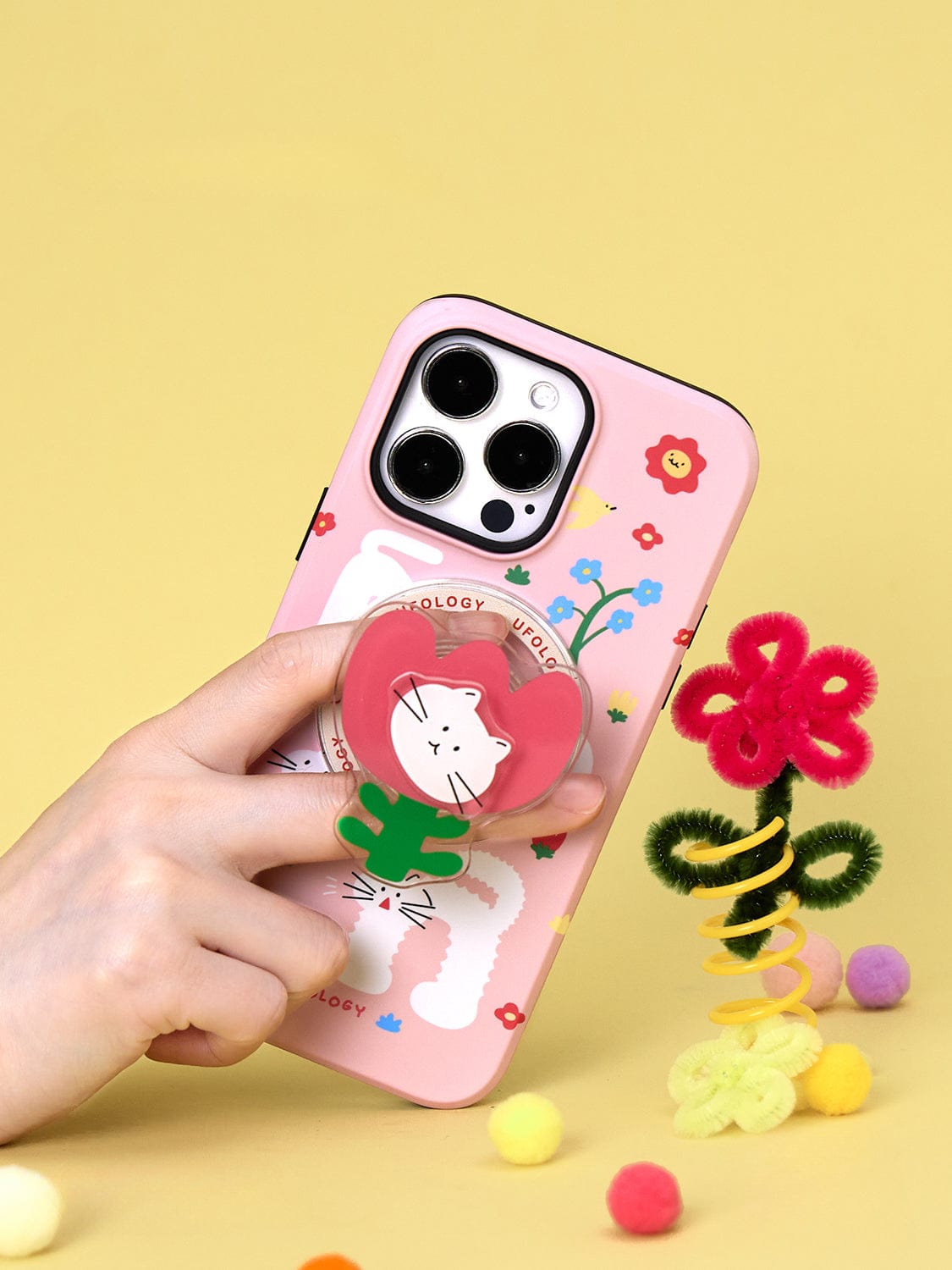 A hand holding the Spring Kitty MagSafe iPhone 16 Pro Max Case, showcasing its playful floral design and cat stand, with colorful pipe cleaner art in the background.