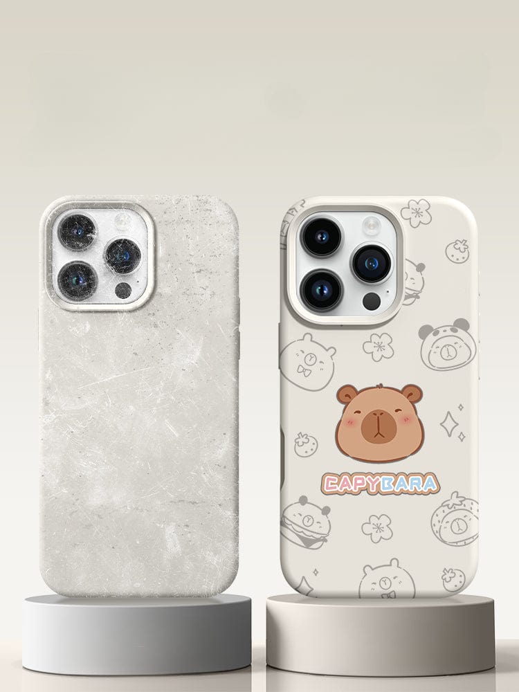 Two smartphones in protective cases on stands. One case is a triple-layer shockproof structure with a marble texture, while the other, an iPhone 16 Pro Max Cute Capybara Silicone Protective Cover with Shockproof Triple-Layer Design and Anti-Fingerprint Coating, features cartoon capybara illustrations and the "CAPYBARA" text. Both phones have triple-lens cameras.