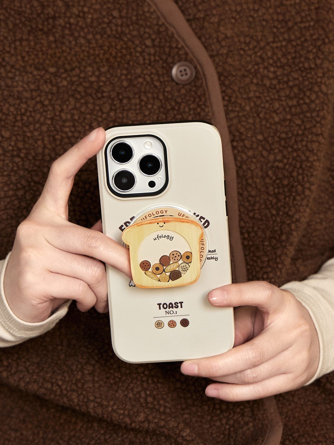 A person wearing a brown sweater holds the Morning Toast MagSafe iPhone 16 Pro Max Case, which has a built-in PopSocket featuring a cartoon slice of toast and the word "Honey" surrounded by cookie illustrations. The beige smartphone case also displays the words "Toast No. 1" at the bottom.