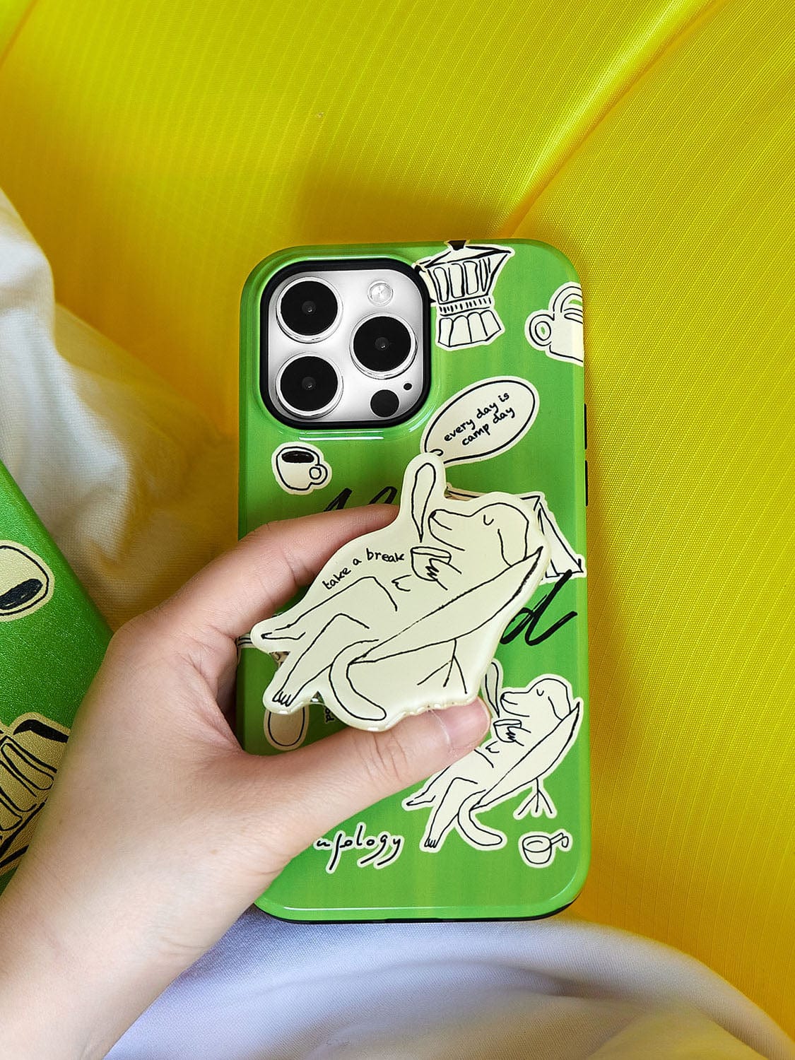 A hand holding a Lazy Day Elephant MagSafe iPhone 16 Pro Max Case, featuring a playful green design with four camera lenses and various stickers including a cat and coffee-related designs.