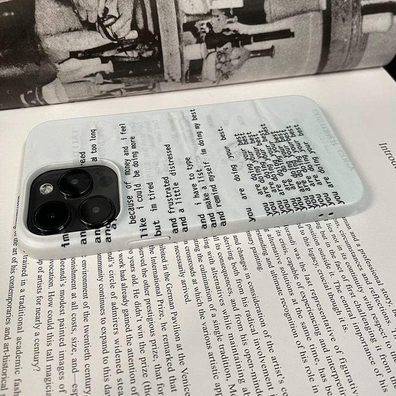 A close-up image of the Affirmation Script iPhone 16 Pro Max Case placed on top of an open book. This minimalist design features transparent sections with printed text that align seamlessly with the book's passages underneath, enhancing its positive vibes. The phone's camera lenses are visible in the upper left corner, contributing to its sleek design.