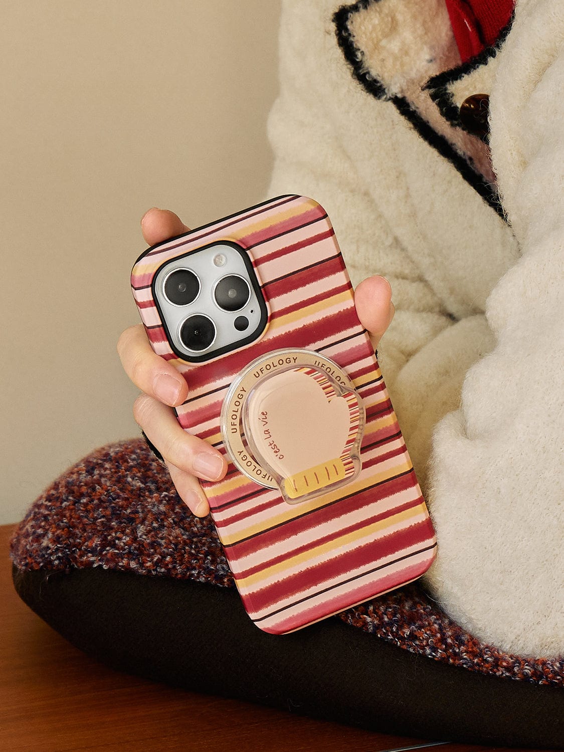 A person wearing a cream-colored fleece jacket with a black and red collar holds an iPhone 16 Pro Max encased in the Autumn Stripes MagSafe iPhone 16 Pro Max Case, featuring shades of red, white, and brown. The case includes a cozy mug design stand with an all-inclusive protective cover that has a round beige grip accessory attached to the back.