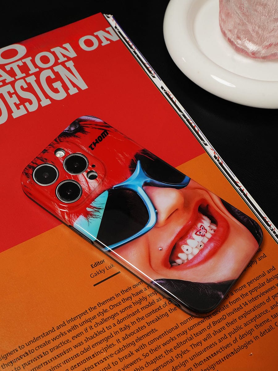 Close-up of the Bold Rockstar iPhone 16 Pro Max Case featuring a vibrant pop art design with a graphic of a smiling face wearing sunglasses and braces. The case is placed on an orange and red book titled "Innovation on Design.