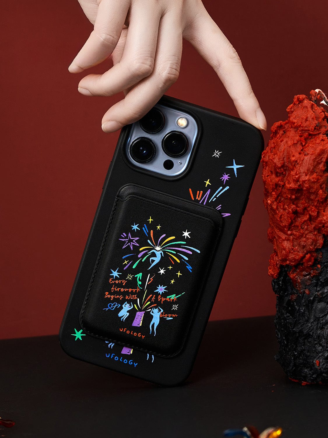 A hand holding a black smartphone with the Firework Fantasy MagSafe iPhone 16 Pro Max Case, which features a vibrant, illustrated design of fireworks and figures on its back. The case includes a special compartment. The background blends deep red and black hues, with a textured object on the right side.