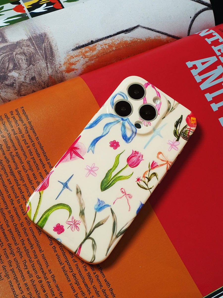 A Floral Dreams iPhone 16 Pro Max Case, featuring an elegant, colorful botanical design, adorns the surface of an open book filled with vibrant pages.