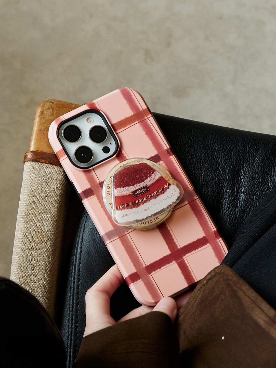 A person is holding a smartphone encased in the Cozy Plaid MagSafe iPhone 16 Pro Max Case, which showcases a pink and red plaid pattern and features a dessert-themed pop socket. The phone rests on a black leather surface, with the case adding an extra touch of style and protection.