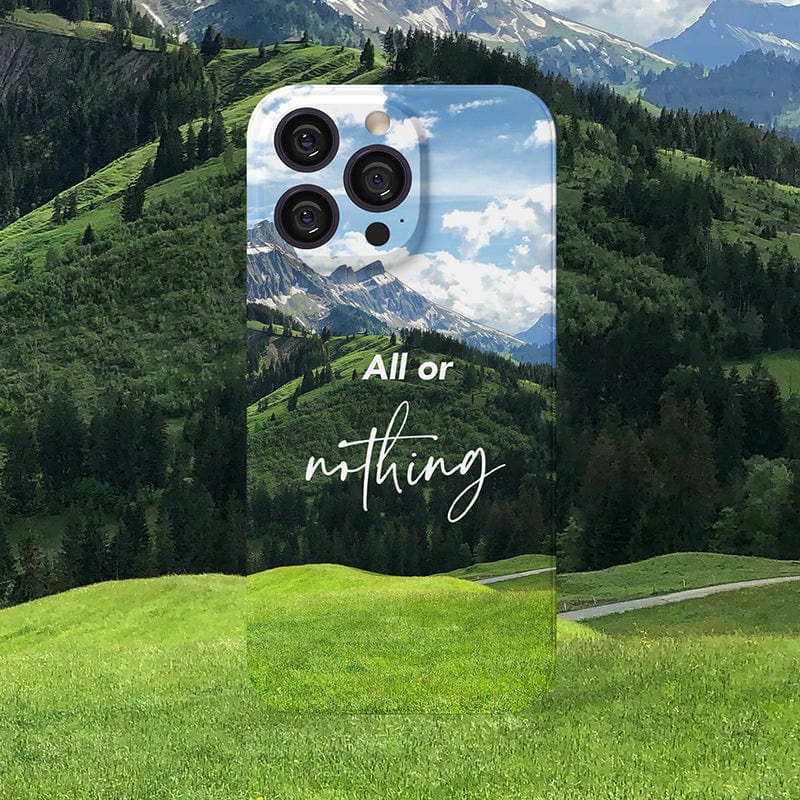 The Mountain Landscape iPhone 16 Pro Max Case - Inspiring Scenic Design with "All or Nothing" Quote is showcased against a backdrop of lush green hills and mountains, featuring a scenic landscape with a blue sky and clouds, along with the inspirational text "All or Nothing" in white handwritten-style font.