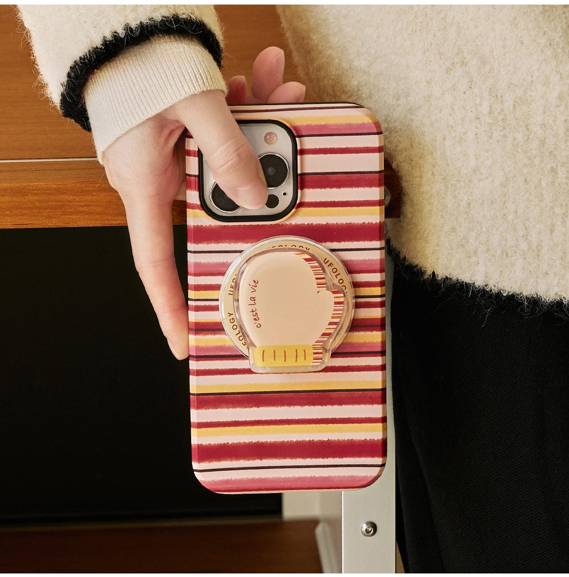 A close-up of a hand holding the Autumn Stripes MagSafe iPhone 16 Pro Max Case, featuring colorful stripes in shades of red, yellow, and white. The case includes a circular grip accessory on the back designed as part of the Cozy Mug collection with a built-in stand. The person is wearing a sweater with cream-colored cuffs.