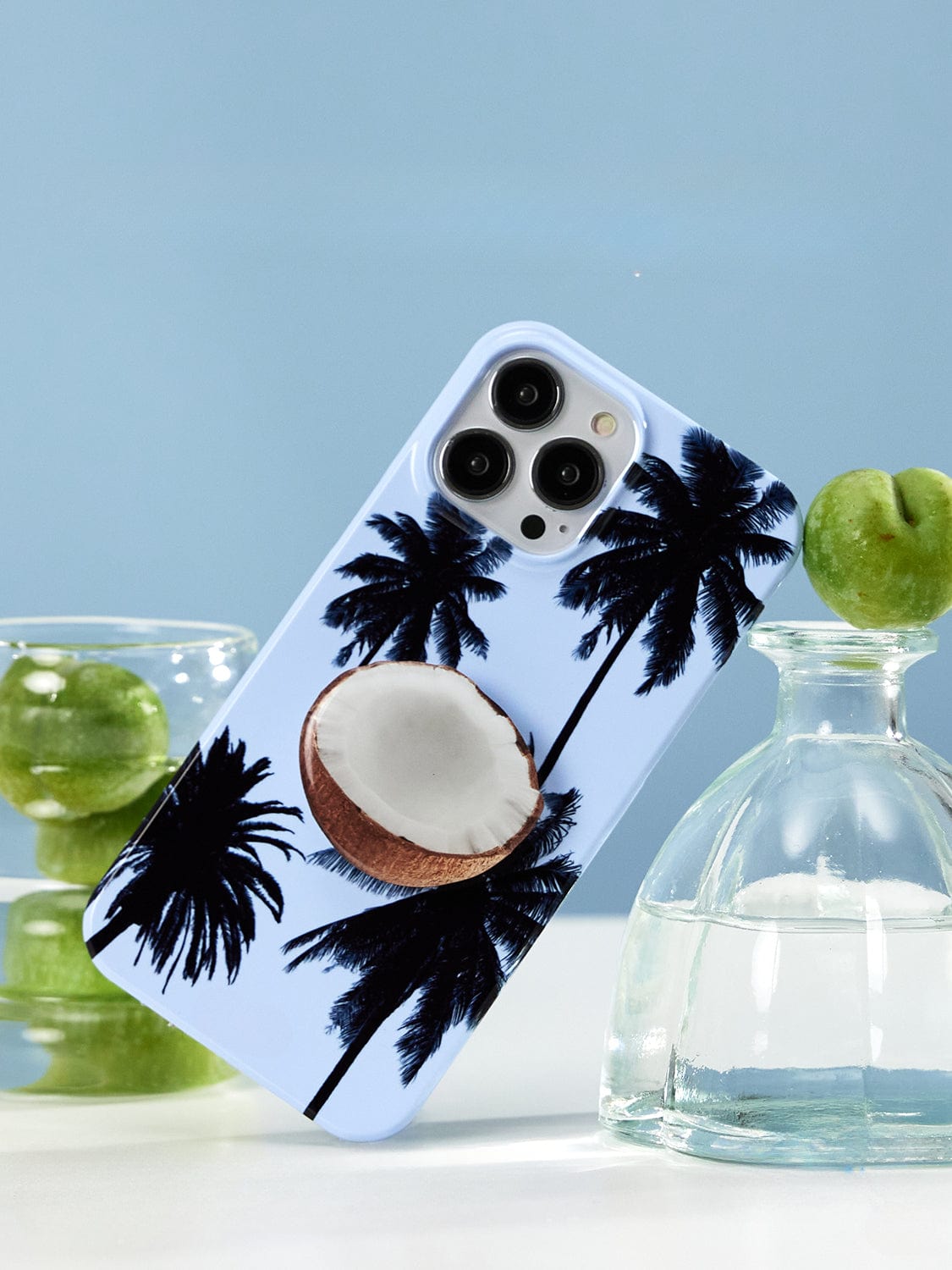 The Tropical Escape MagSafe iPhone 16 Pro Max Case, showcasing a palm tree design with a halved coconut stand, is propped up vertically. In the background, two green apples—a jarred one and another placed on a glass bottle—sit against a light blue backdrop.